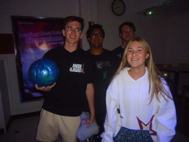 Four members at a bowling alley, one holding a ball beside a champions poster